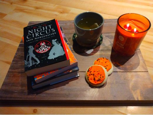 The Night Circus by Erin Morgenstern sitting on a tray with tea, a cozy candle, and some Halloween cookies.