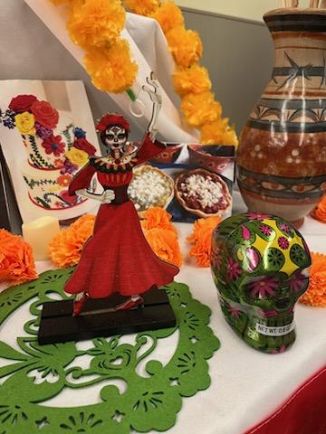 A photo taken by Cynthia Guevara on 10/30/23 of a Dia de los Muertos altar, with a traditional Mexico flowers, Cempasúchil (marigolds), a calavera (skull), a picture of picadas, which are a traditional Mexican food and a cardboard cut out of La Catrina, a symbol of the day of the dead.